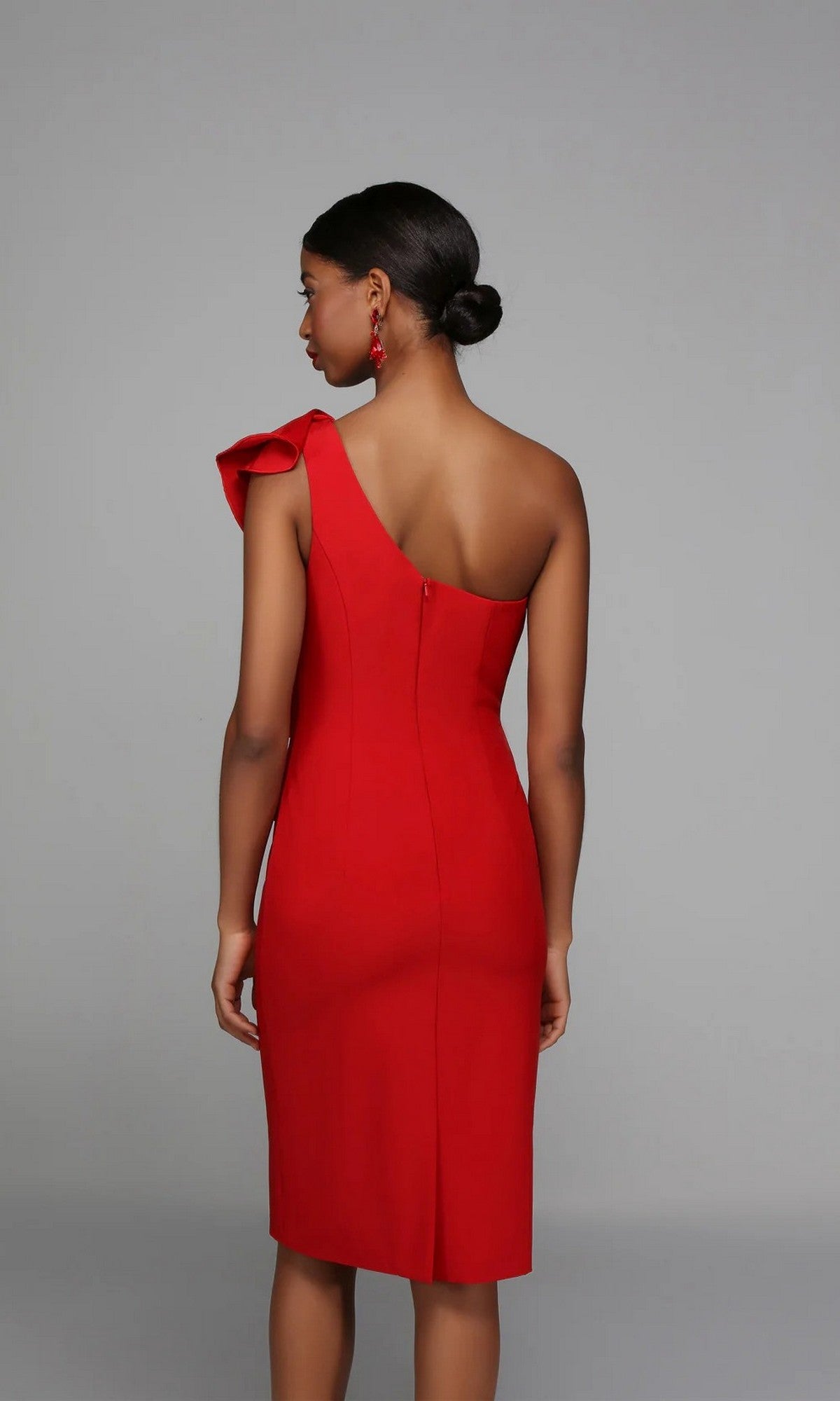 A woman wearing the Alyce 70008 knee-length semi-formal dress with a fitted silhouette and one-shoulder ruffle stands against a plain gray background, facing away from the camera.
